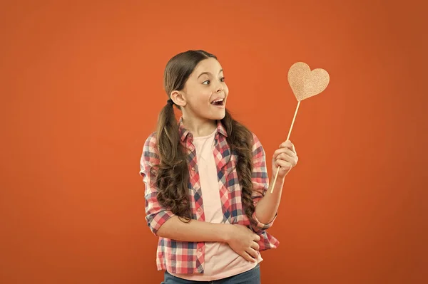 I have my valentines. Adorable valentines girl on orange background. Little child looking at prop heart on valentines day. Cute kid holding fancy valentines day party props on stick — Stock Photo, Image