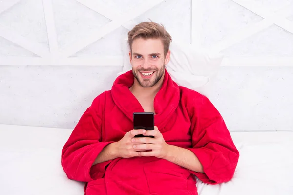 Living a mobile lifestyle. Cheerful guy using mobile device in bed. Handsome man smiling with mobile phone in hands. Mobile communication and wireless technologies in home