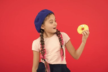 Pişmiş Donuts büyük fanlar çocuklar. Resist taze yapılmış çörek için imkansız. Kız tatlı çörek elinde kırmızı camlı arka plan tutun. Çocuk gülümseyen kız donut yemeye hazır. Şeker dükkanı ve ekmek kavramı