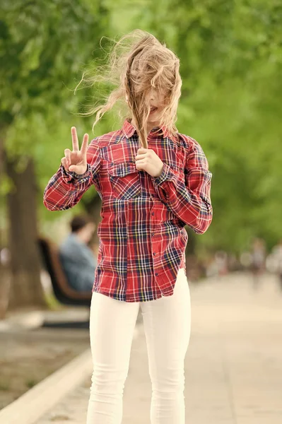 Het is winderig buiten. Klein kind met rommelig haar op zomerdag. Leuk kind geniet van haar haar zwaaiende en toont overwinnings gebaar. Schattig klein meisje met lange blonde haren waait in de wind. Haarverzorging — Stockfoto