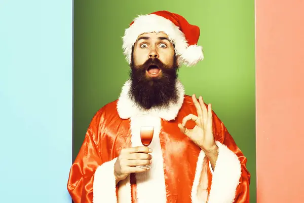 Bonito barbudo santa claus homem com longa barba no rosto surpreso segurando vidro de tiro alcoólico no vermelho natal ou xmas casaco e chapéu de ano novo no colorido estúdio backgroun — Fotografia de Stock