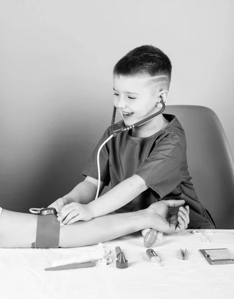 Αγόρι χαριτωμένο παιδί μέλλων γιατρός καριέρας. Υγειονομική περίθαλψη. Παιδί μικρό γιατρό κάθονται τραπέζι με στηθοσκόπιο και ιατρικά εργαλεία. Η ιδέα της ιατρικής. Μετρώντας την αρτηριακή πίεση. Ιατρική εξέταση. Ιατρική εκπαίδευση — Φωτογραφία Αρχείου