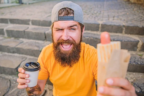 Um tipo a comer cachorros quentes. Homem barbudo comer salsicha saborosa e beber copo de papel. Conceito de comida de rua. Nutrição de estilo de vida urbano. Comida de plástico. hipster despreocupado comer junk food while sit stairs. Lanche de bom humor — Fotografia de Stock