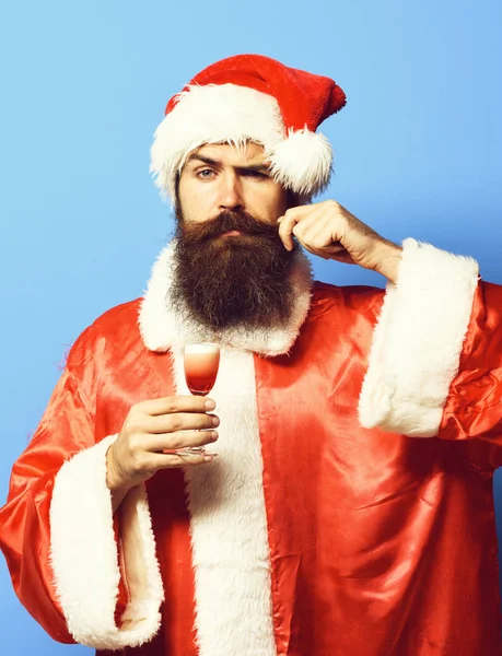 Bonito barbudo santa claus homem com longa barba no rosto sério segurando vidro de tiro alcoólico no Natal ou xmas suéter e chapéu de ano novo e tocando bigode no estúdio azul backgroun — Fotografia de Stock