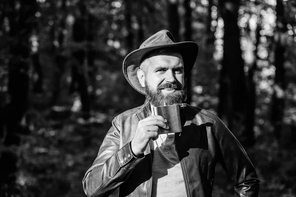 man hipster relax in autumn forest. Spring sunny weather. camping and hiking. mature male with brutal look drink alcohol from metallic flask. Bearded man in cowboy hat in park outdoor. Smiling cowboy