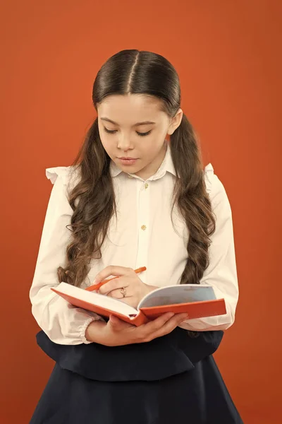 Colegiala escribir notas sobre fondo naranja. lección de lectura. obtener información libro de formularios. de vuelta a la escuela. niño pequeño concentrado en el trabajo. Niña con uniforme escolar. escritura en el libro de trabajo — Foto de Stock