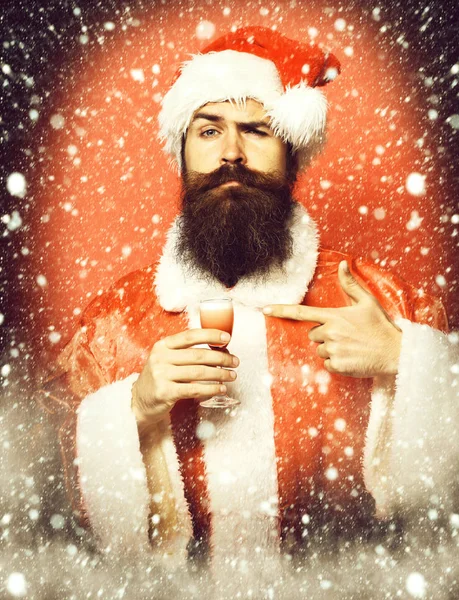 Guapo barbudo hombre de Santa Claus con barba larga en la cara seria sosteniendo vaso de trago alcohólico en navidad o suéter de Navidad y sombrero de año nuevo y se muestra en él en el fondo del estudio rojo —  Fotos de Stock