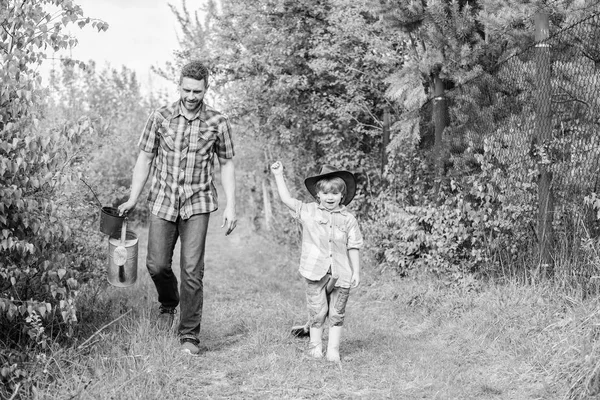 仕事の準備ができました。牧場でカウボーイハットをかぶった父と息子。エコファーム小さな男の子の子供は、農業で父親を助けます。水やり缶とポットを使用してください。庭の設備。幸せな地球の日。家族の木の看護 — ストック写真