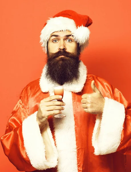 Guapo barbudo hombre de Santa Claus con barba larga en la cara seria sosteniendo vaso de trago alcohólico en navidad o suéter de Navidad y sombrero de año nuevo que muestra fresco en el fondo del estudio rojo —  Fotos de Stock