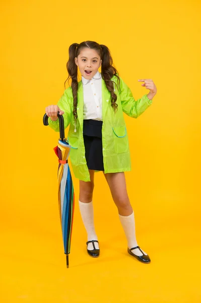 Ela é uma vítima de moda. Escolar bonito apontando dedo com olhar de moda no fundo amarelo. Criança em capa de chuva segurando guarda-chuva de moda. Roupa de chuva e moda dia chuvoso — Fotografia de Stock