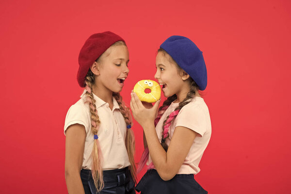 Sweets shop and bakery concept. Kids huge fans of baked donuts. Share sweet donut. Girls in beret hats hold glazed donut red background. Kids playful girls ready eat donut. Friendship and generosity