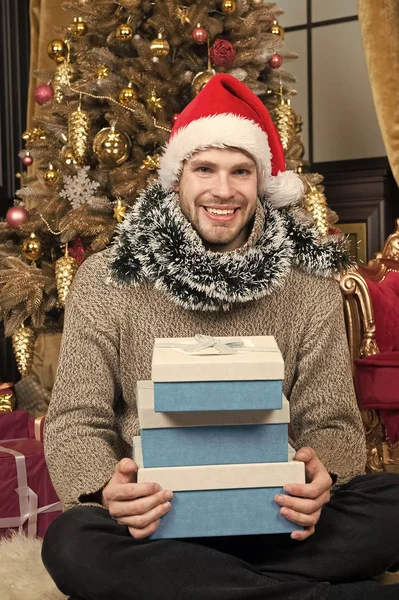 Christmas time. Man in santa hat hold xmas presents. Happy man with xmas gift boxes. Guy is celebrating Christmas at home. Christmas Eve. Delivery Christmas gifts — Stock Photo, Image