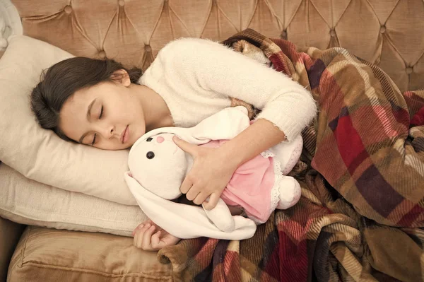 Duerme con juguete. Chica disfrutar del tiempo de la noche con juguete favorito. Niño acostado cama y abrazo conejito juguete sofá almohada manta fondo vista superior. Niña niña usar pijama abrazo conejito. Juega juguete suave antes de ir a dormir —  Fotos de Stock