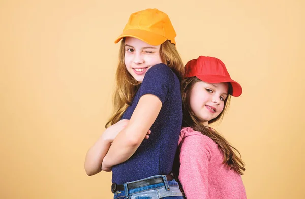 Cool girls. Sisters stand back to back beige background. Little cute girls wearing bright baseball caps. Modern fashion. Hats and caps. Stylish accessory. Kids fashion. Feeling confident wearing caps