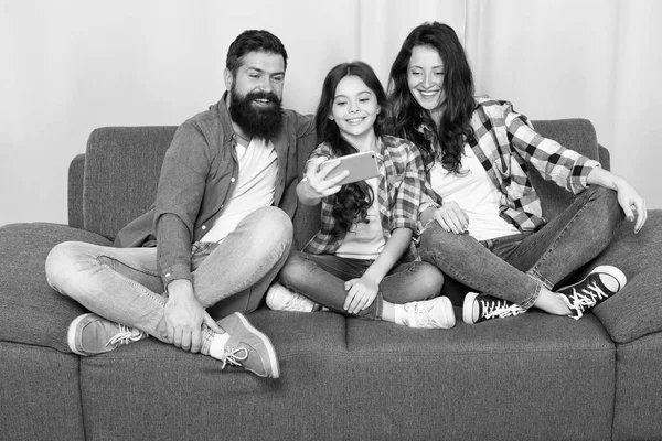 Capture happy moments. Family spend weekend together. Use smartphone for selfie. Friendly family having fun together. Mom dad and daughter relaxing on couch. Family posing for photo. Family selfie