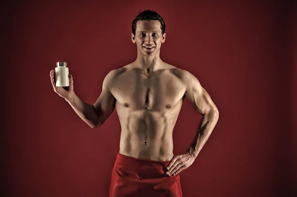 Man with strong belly hold pill jar. Red. Presenting product. Power and energy. Fitness and dieting concept. Sexy muscular man on red background. Anabolics and drugs for sport