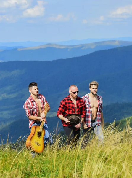 Group of young people in checkered shirts walking together on top of mountain. Men with guitar hiking on sunny day. Tourists hiking concept. Hiking with friends. Enjoying freedom together. Long route