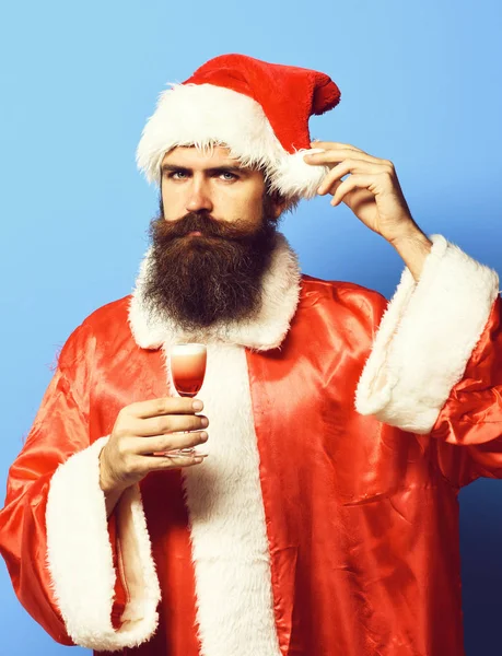 Bonito barbudo santa claus homem com longa barba no rosto sério segurando vidro de tiro alcoólico no Natal ou xmas suéter e chapéu de ano novo e tocando pompon no estúdio azul backgroun — Fotografia de Stock