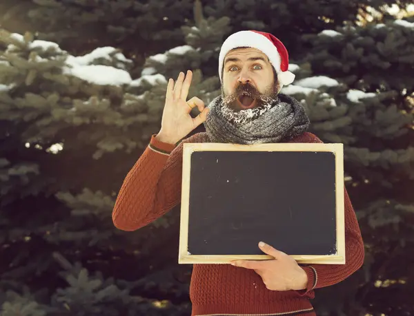 Noel Baba şapkalı heyecanlı adam, beyaz don ile kaplı sakallı ve bıyıklı hipster, boş siyah tahta veya tebeşir le doğal arka planda kış gününde ok jest verir, kopya spa — Stok fotoğraf