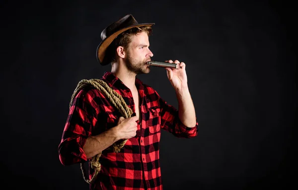 Sheriff concept. Brutal cowboy drinking alcohol. Western culture. Man handsome unshaven cowboy black background. Western life. Man wearing hat hold rope and flask. Lasso tool of American cowboy