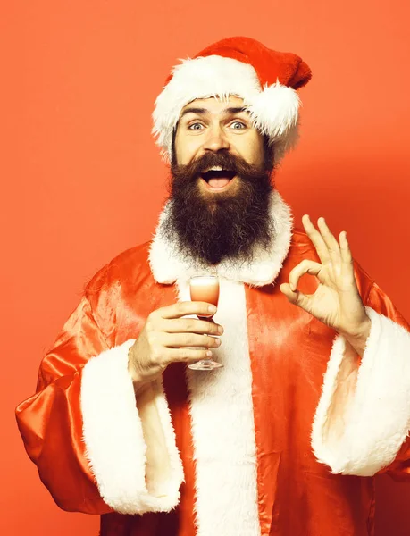 Bonito barbudo santa claus homem com longa barba no sorriso rosto segurando vidro de tiro alcoólico no Natal ou xmas suéter e chapéu de ano novo e mostrando ok no estúdio vermelho backgroun — Fotografia de Stock