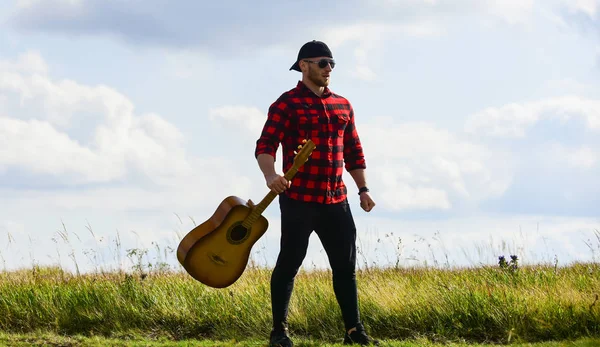 Country style. Summer vacation. Hiking song. Inspired country musician. United with nature. Handsome man with guitar. Country music concept. Guitarist country singer stand in field sky background