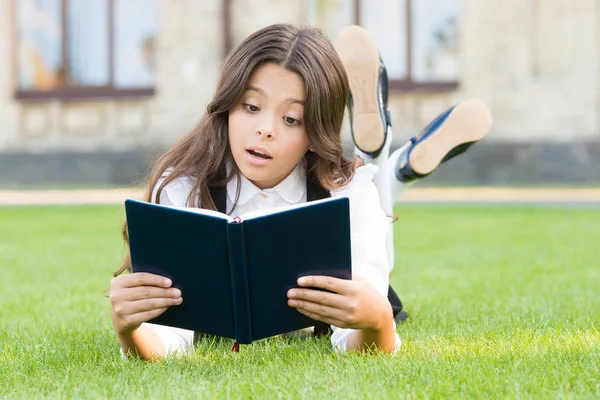 Studuji koncept. Mimoškolní čtení. Roztomilé malé dítě čtení knihy venku. Základní vzdělání. Roztomilá holčička se učí číst. Školačka školní uniforma ležící na trávníku s oblíbenou knihou — Stock fotografie