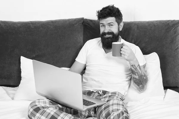 早晨从咖啡和互联网开始。生意人在床上留胡子的男人在笔记本电脑上工作，喝咖啡。成熟的男性与胡子在床上的睡衣。精力和疲劳。有电脑的商人 — 图库照片
