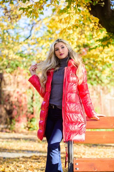 Lady attractive posing in jacket near bench. Woman fashionable blonde with makeup stand in autumnal park. Jacket for fall season concept. Girl wear red bright warm jacket. Fall fashion concept