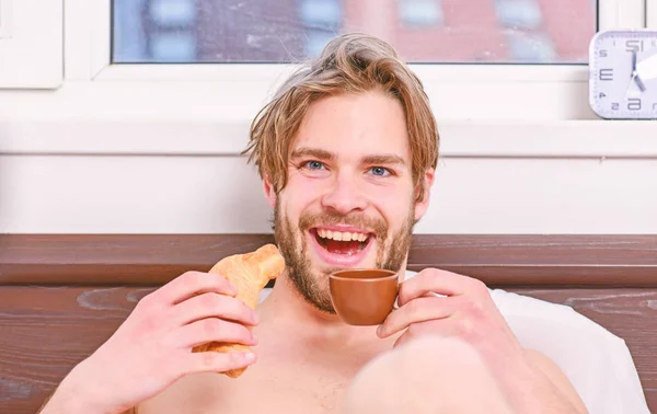 French breakfast stereotype. Sleeping man being woken by mobile phone in bedroom. Guy having coffee and holds croissant while lay bed in bedroom or hotel room. Breakfast in bed. — Stock Photo, Image