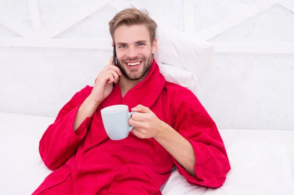 Break time. Happy guy in bathrobe relaxing during break in bed. Handsome man talking on mobile phone during coffee break in bedroom. Enjoying after bath spa break