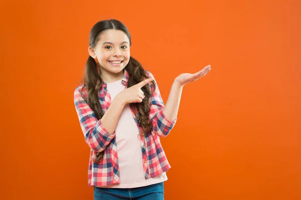 Look here. happy child. happy childrens day. childhood memory. small girl orange background. kid fashion. smiling school girl. kid long hair presenting product. small girl checkered shirt. copy space — Stock Photo, Image