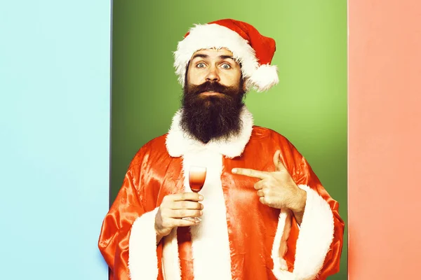 Knappe bebaarde Kerstman man met lange baard op grappig gezicht houden van glas alcoholische shot in rode kerst of Xmas jas en Nieuwjaar hoed op kleurrijke Studio backgroun — Stockfoto