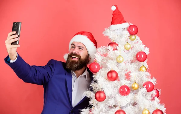 Capturer des souvenirs de Noël. Homme à la barbe et au visage joyeux, saluez Noël. Concept vacances d'hiver. Santa claus prend selfie près de l'arbre de Noël sur fond rouge. Père Noël détient téléphone portable — Photo