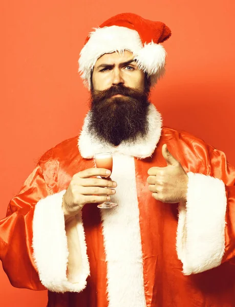 Knappe bebaarde Kerstman man met lange baard op ernstige gezicht houden van glas alcoholische shot in Kerstmis of Xmas trui en Nieuwjaar hoed weergegeven cool op rode studio backgroun — Stockfoto
