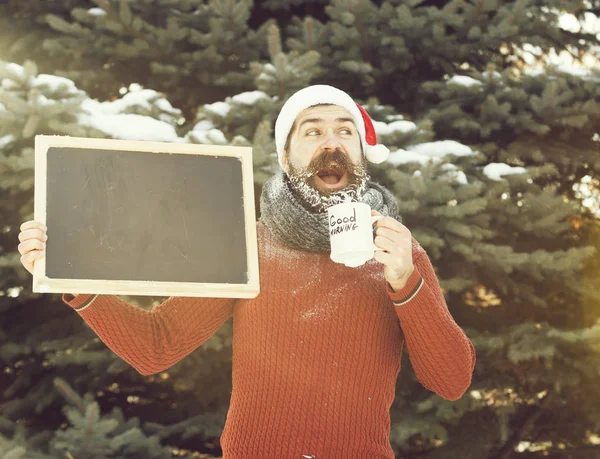 Noel Baba şapka yakışıklı mutlu adam, sakal ve bıyık beyaz don ile kaplı sakallı hipster, doğal arka plan üzerinde açık havada boş siyah tahta ve kahve fincanı kış günü tutar, kopya spa — Stok fotoğraf
