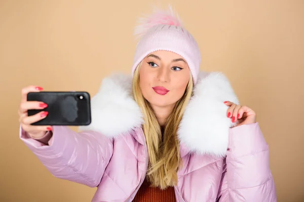 woman in beanie hat. happy winter holidays. web blogger. girl in puffed coat make selfie. faux fur fashion. warm winter clothing. phone selfie. flu and cold season. Leather bag fashion