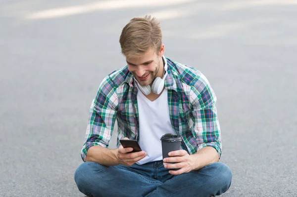 guy drink coffee outdoor. man sit on ground. carefree student in headset use phone. online education. listen music. ebook concept. man checkered shirt. student relax and recharge. coffee to go