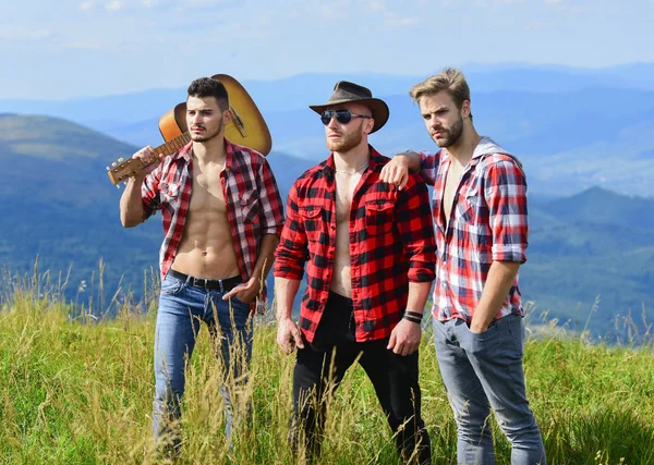 Men with guitar hiking on sunny day. Group of young people in checkered shirts walking together on top of mountain. Long route. Adventurers squad. Tourists hiking concept. Hiking with friends