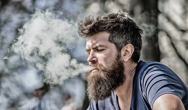Мужчина с длинной бородой, расслабленный от курения. Человек с бородой выдыхает дым. Облака ароматизированного дыма. Концепция снятия стресса. Бородатый человек курит вапе. Курение электронных сигарет. Курящее устройство — стоковое фото