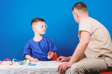 Tatlı çocuk ve babası doktor. Hastane çalışanı. Sağlık hizmetleri. Tıp konsepti. Küçük doktor, masada tıbbi aletlerle oturuyor. Hastalık tedavisi. Baba ve oğul tıp hanedanlığı. Tıbbi muayene
