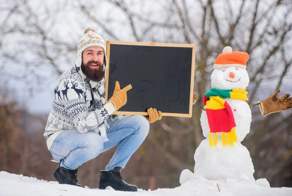 Upcoming event. Man with beard hold chalkboard copy space. Winter announcement. Hipster knitted hat and gloves show blackboard. Guy and snowman snowy nature background. Winter event. Winter holidays