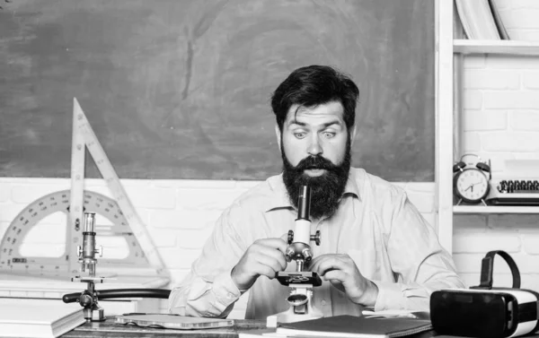 School teacher looking microscope. Fascinating research. Set up microscope. Teacher sit desk with microscope. Man bearded hipster classroom chalkboard background busy with biological observation