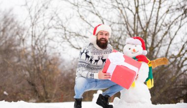 Adam gülen surat, karlı doğa geçmişi. Sakallı hippi hediye kutusu. Sürpriz konsept. Kış oyunları. Kış aktivitesi. Kış tatili Adam kardan adam yaptı. Noel Baba şapkası dışarıda eğleniyor.