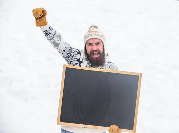 Winter event. Man with beard hold chalkboard copy space. Check it out. Winter holidays. Emotional guy snowy nature background. Winter announcement. Hipster knitted hat and gloves show blackboard — Stock Photo, Image