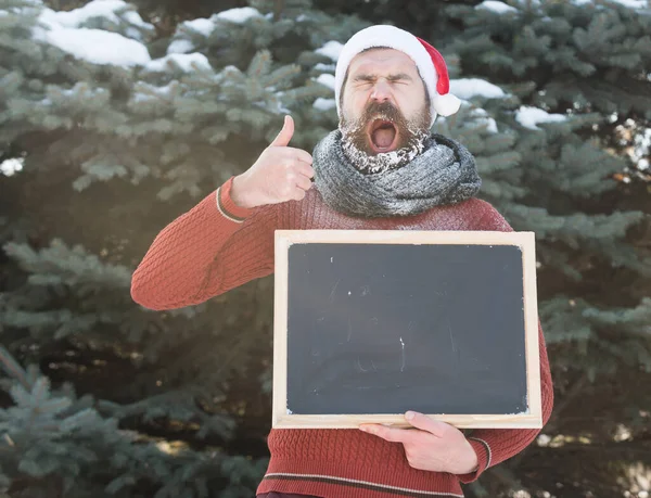 Schreeuwende man in kerstman hoed, hipster met baard en snor bedekt met witte vorst, met blanco zwart bord of schoolbord geeft duimen omhoog gebaar op winterdag op natuurlijke achtergrond, kopiëren s s — Stockfoto