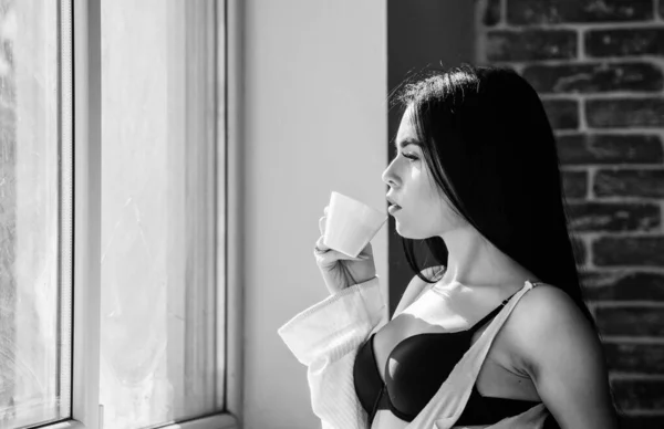 Aantrekkelijke vrouwelijke lingerie ontspannen thuis. Perfecte koffie tijd. Maar eerste koffie. Rust en vrede. SIP van romige cappuccino. Ochtendritueel. Meisje sexy borsten drinken koffie in de buurt van vensterbank — Stockfoto