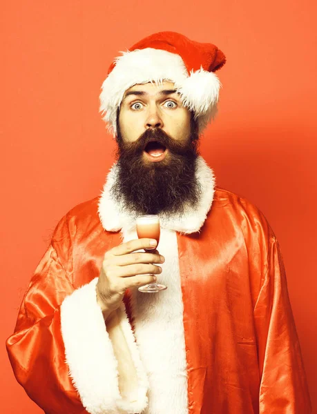 Stilig skäggig jultomte man med långt skägg på överraskat ansikte innehav glas alkoholhaltiga skott i jul eller jul tröja och nyår hatt på röd studio backgroun — Stockfoto