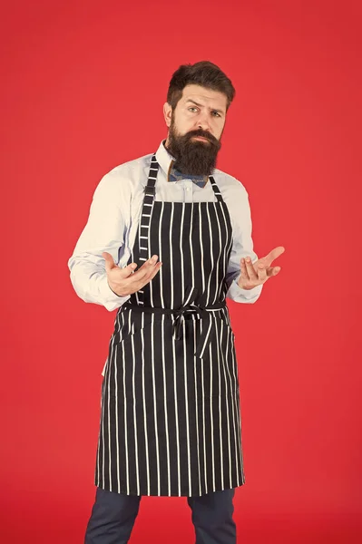 Comment puis-je vous aider ? Homme avec barbe cuisinier tablier hipster. Hipster chef cuisinier fond rouge. Un homme barbu cuisinier. Service et personnel du restaurant. Serveur bien entretenu au travail. Concept de café Hipster — Photo