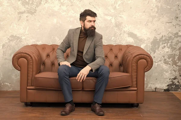 Realtor and rental service. Rent apartment. Bearded man with confident face sit leather couch. Loft interior apartment. Businessman realtor work. Furniture shop. Hipster realtor loft style apartment